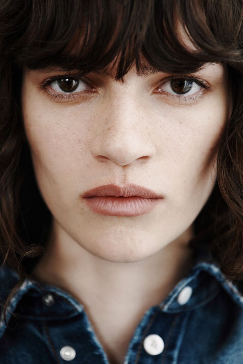 Close-up portrait of a woman with dark wavy hair and fringe bangs wearing a collared blue denim shirt.