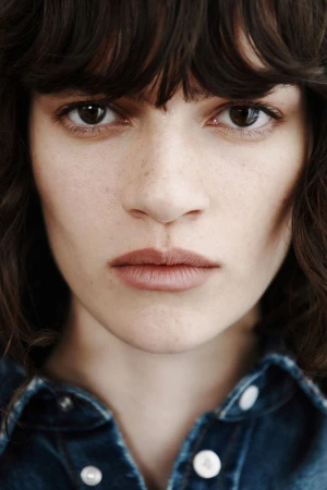 Close-up portrait of a woman with dark wavy hair and fringe bangs wearing a collared blue denim shirt.