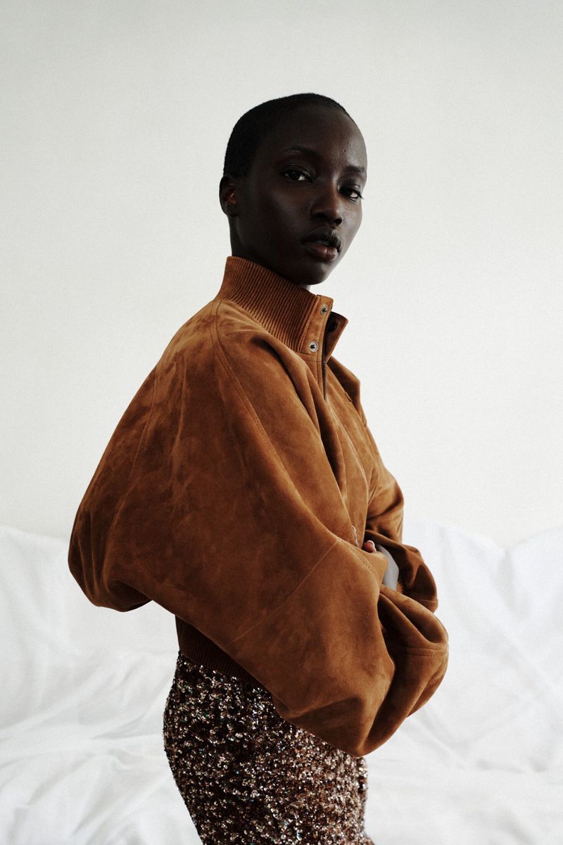 Model Anok Yai posing in a voluminous brown suede jacket and sequined skirt against a draped white background.