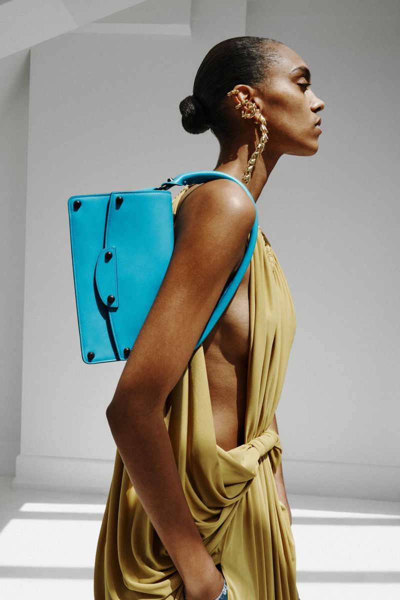 Model in profile wearing a draped mustard backless gown with a bright cyan blue shoulder bag against a white wall.