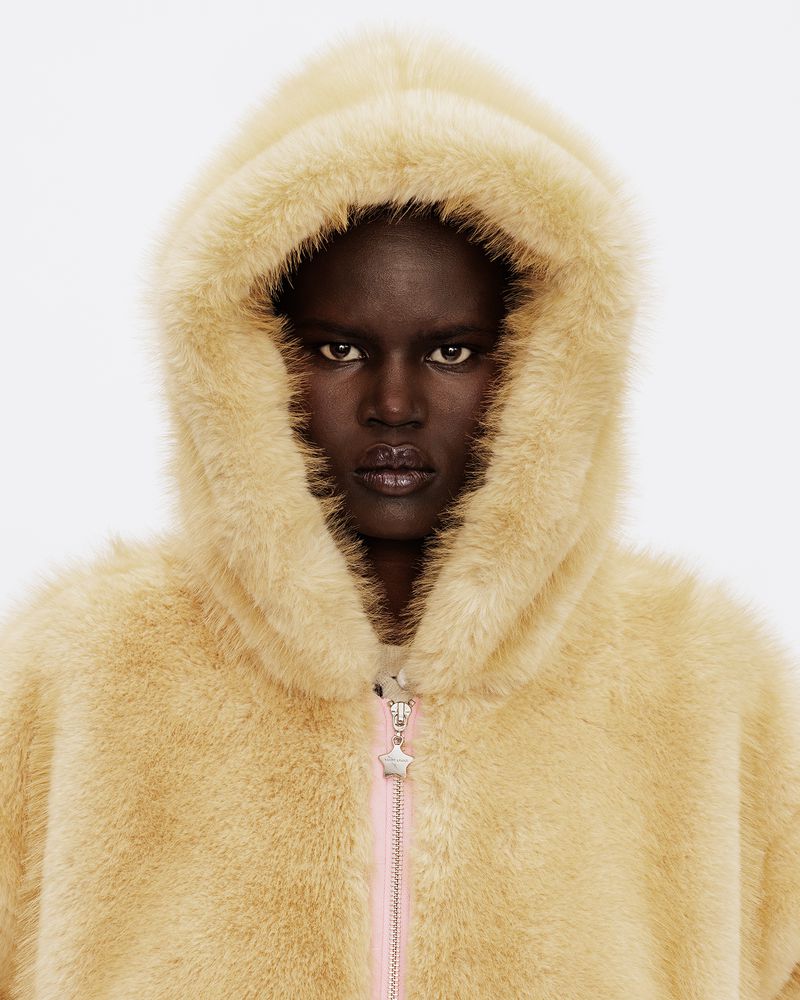 Close-up portrait of a Black woman wearing a pale yellow faux-fur hooded jacket with a star-shaped zipper pull against a white background.