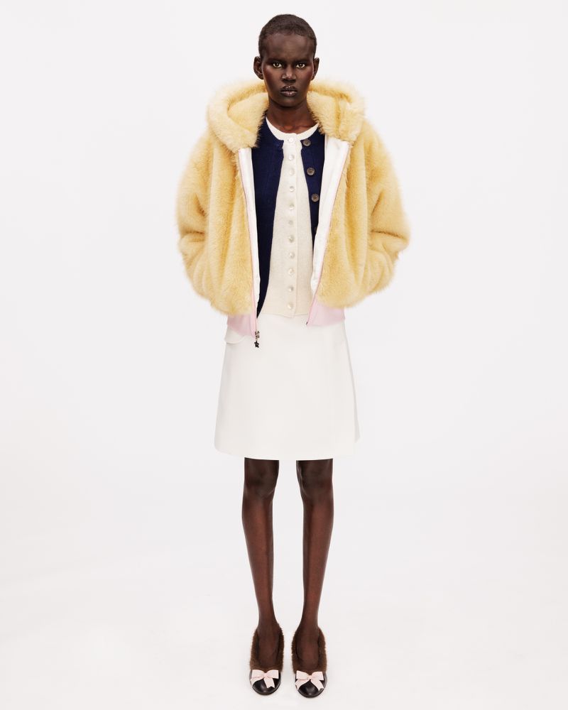 Model wearing a hooded pale yellow fleece jacket, white skirt, and bow-embellished ballet flats on a white seamless backdrop.
