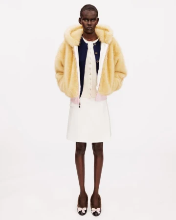 Model wearing a hooded pale yellow fleece jacket, white skirt, and bow-embellished ballet flats on a white seamless backdrop.