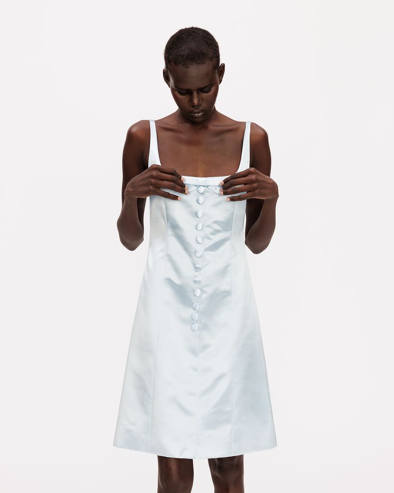 Black woman with buzzcut hair adjusting a pale blue satin sleeveless button-front dress against a plain white studio background.