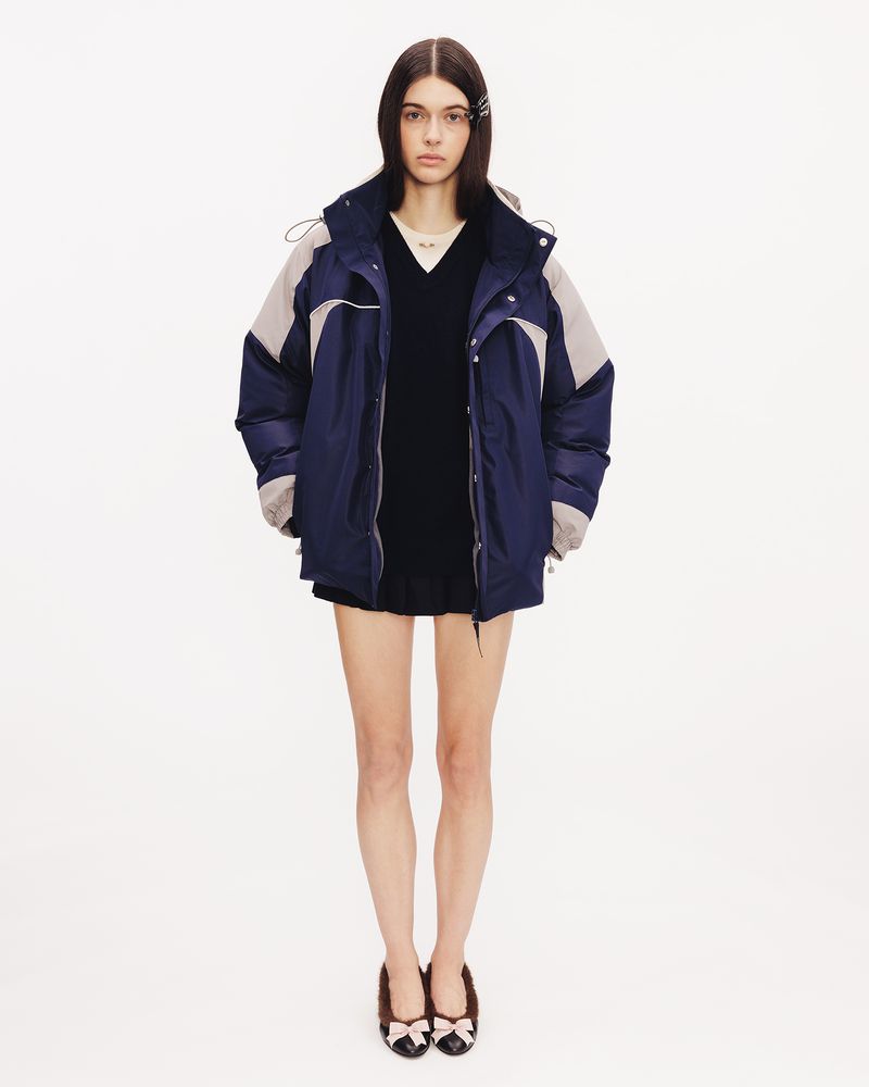 Model wearing an oversized navy blue and grey hooded puffer jacket over a black dress and bow-accented flats on a white seamless backdrop.