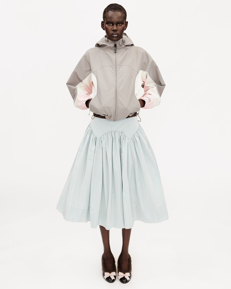 Model wearing a grey color-blocked hooded jacket and pale blue pleated midi skirt standing against a white seamless background.