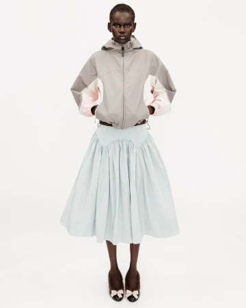 Model wearing a grey color-blocked hooded jacket and pale blue pleated midi skirt standing against a white seamless background.