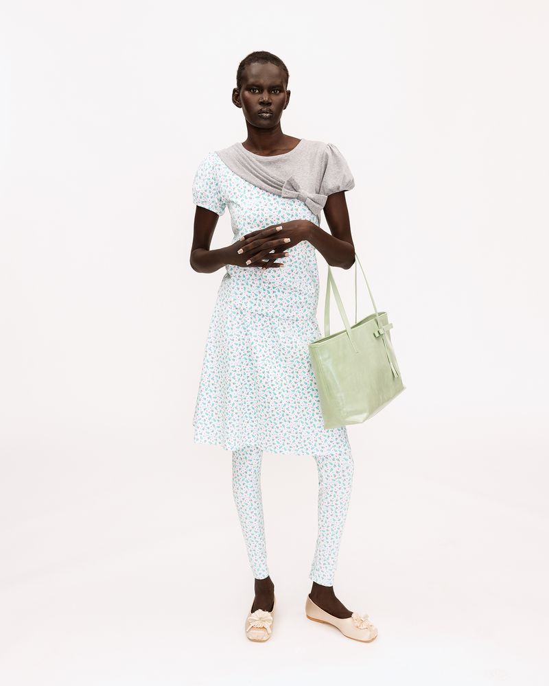 Black female model posing in a blue floral dress, matching printed leggings, grey shrug, and light green tote on a white background.
