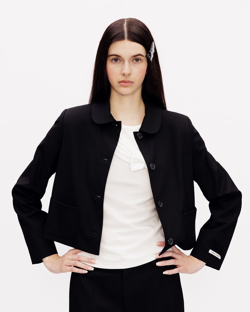Model wearing a black cropped jacket with a rounded collar and white top against a white backdrop.