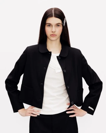 Model wearing a black cropped jacket with a rounded collar and white top against a white backdrop.
