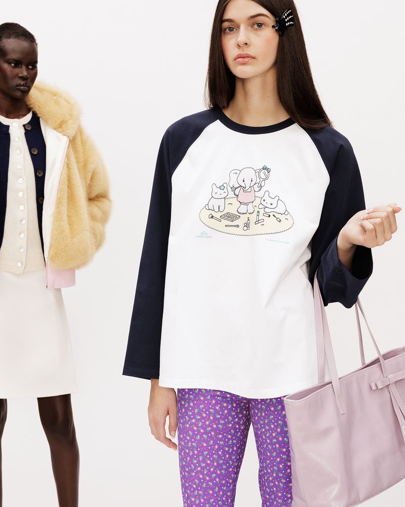 Two female models posing against a white background, wearing graphic raglan top and purple printed pants with a pink tote bag.
