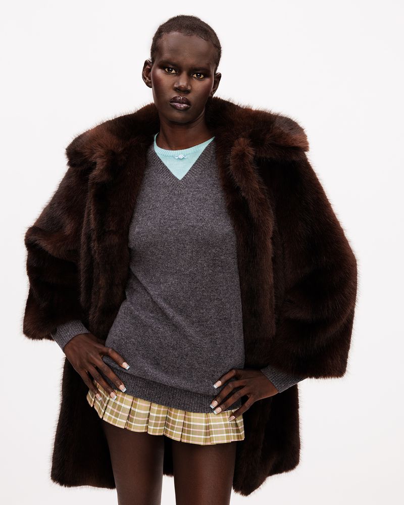 Model posing in an oversized dark brown fur coat, grey v-neck knit sweater, and plaid pleated micro mini skirt on a white background.