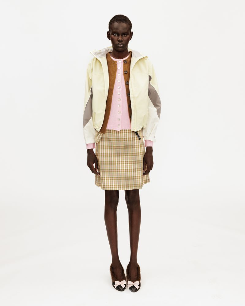 Model wearing a light hooded jacket, pink cardigan, beige plaid mini skirt, and bow flats against a white seamless background.