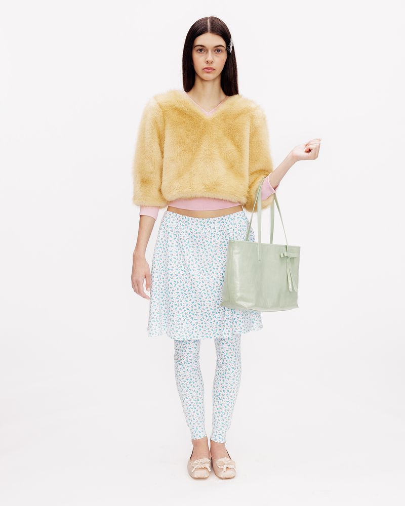 Model wearing a yellow cropped plush sweater, matching floral skirt and tights, holding a green tote against a white backdrop.