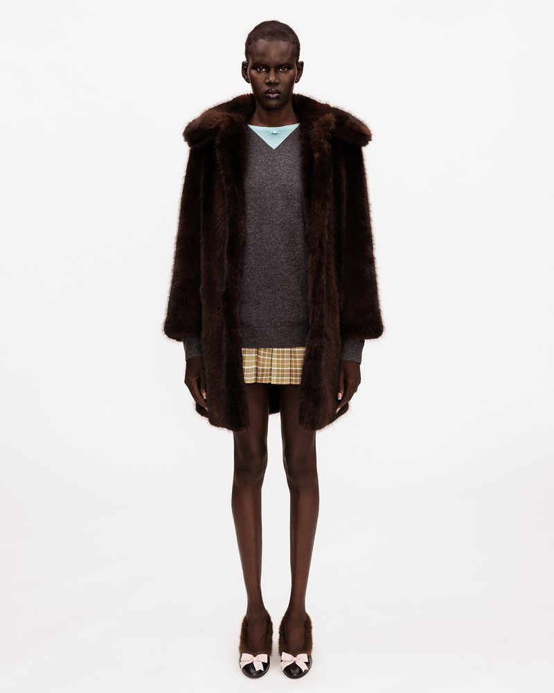 A Black female model in a dark brown fur coat, grey knit sweater, and plaid mini skirt standing against a white seamless backdrop.