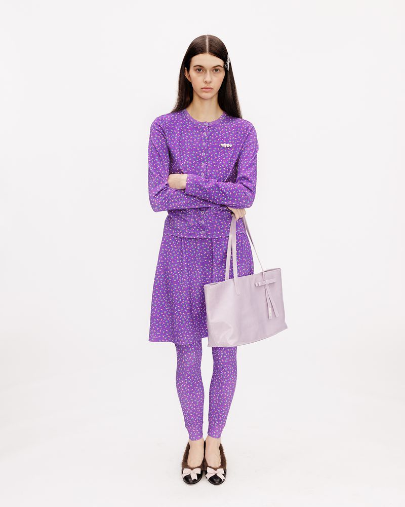 Model wearing a purple printed cardigan, skirt, and leggings set holding a lilac tote bag against a white studio background.