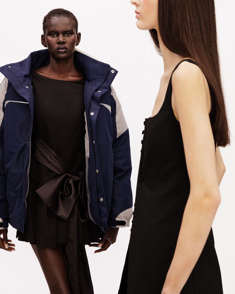 Two models posing in studio, one in a navy blue oversized jacket and black bow dress, another in profile wearing a black sleeveless dress.