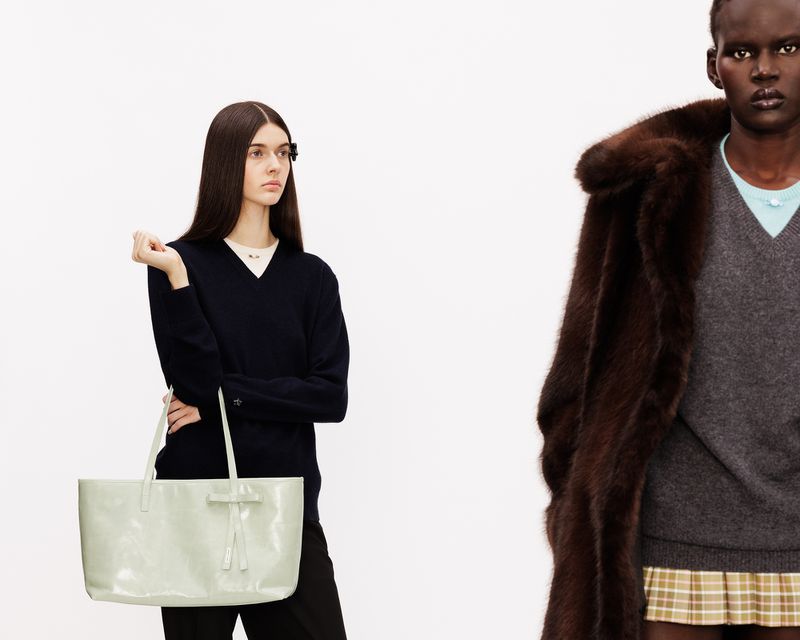 Two models posing in studio against a white background, one holding a pale mint tote bag and the other wearing a dark brown fur coat.