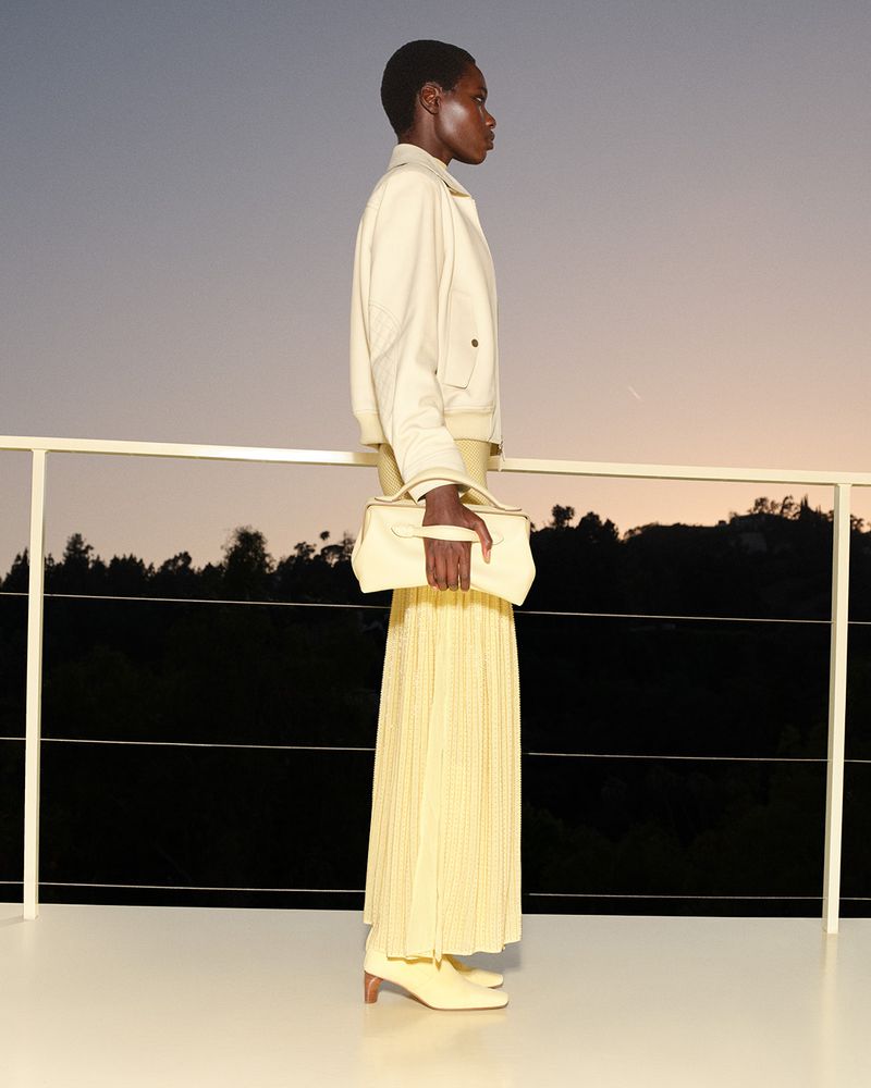 Model in light-colored jacket and pleated skirt holding a handbag on a balcony against a sunset sky.
