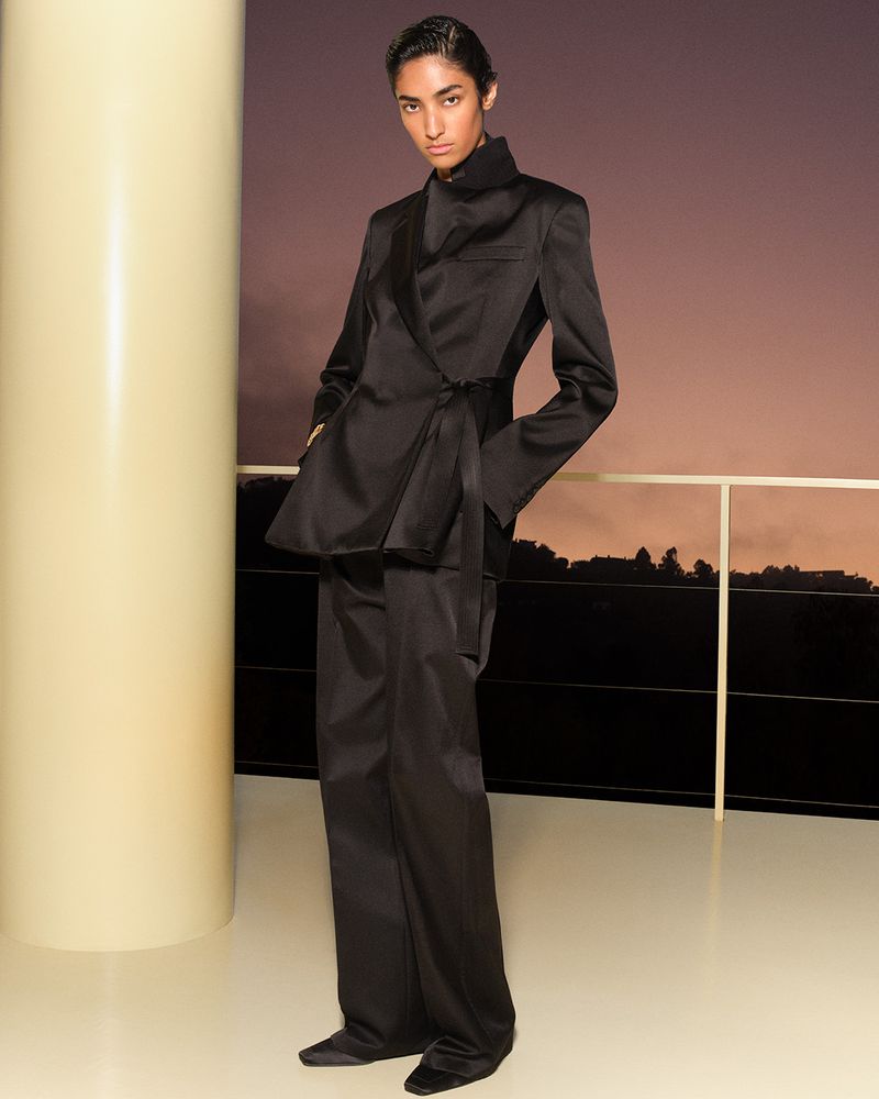 Model wearing a structured black suit standing on a balcony at sunset with a large cream column in the foreground