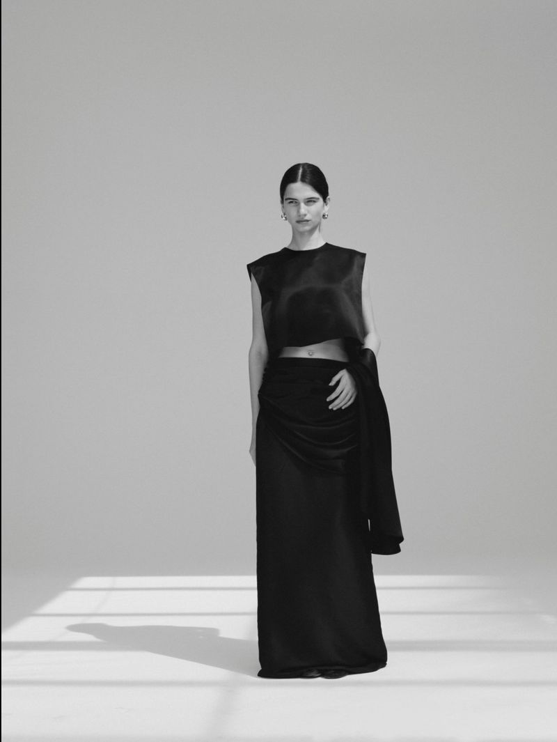 Model Peyton Cole standing in a black satin crop top and long skirt in a studio with floor shadows.