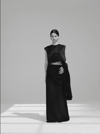 Model Peyton Cole standing in a black satin crop top and long skirt in a studio with floor shadows.