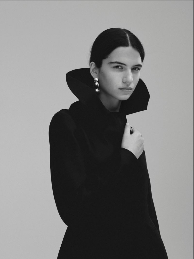 Model Peyton Cole in a high structured collar black coat with a metallic drop earring against a neutral backdrop.