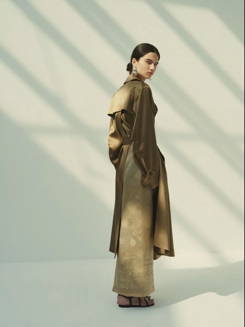 Model Peyton Cole looking over her shoulder wearing a satin olive trench coat and long golden skirt in a bright studio.