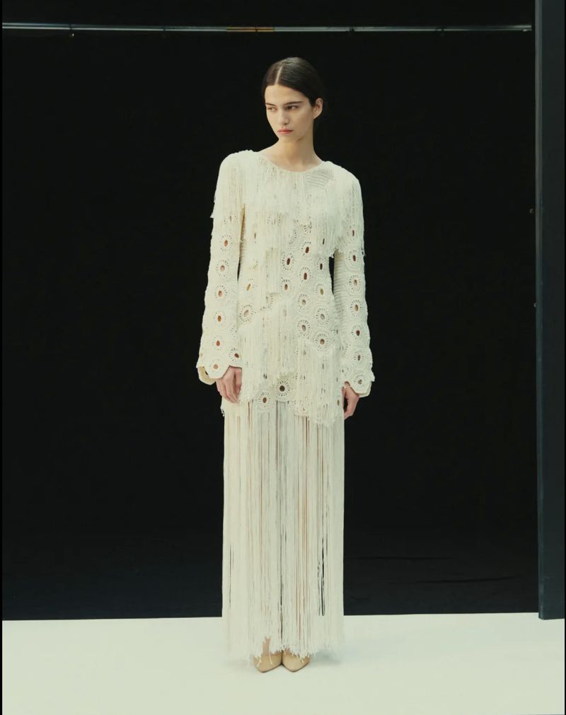 Model Peyton Cole poses standing in an ivory fringed knit maxi dress against a black studio backdrop with a white floor.