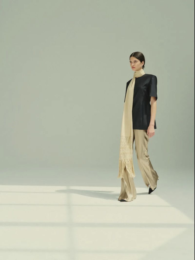 Peyton Cole walks in a dark short-sleeved top, long fringed cream scarf, and champagne trousers against a pale green studio backdrop.