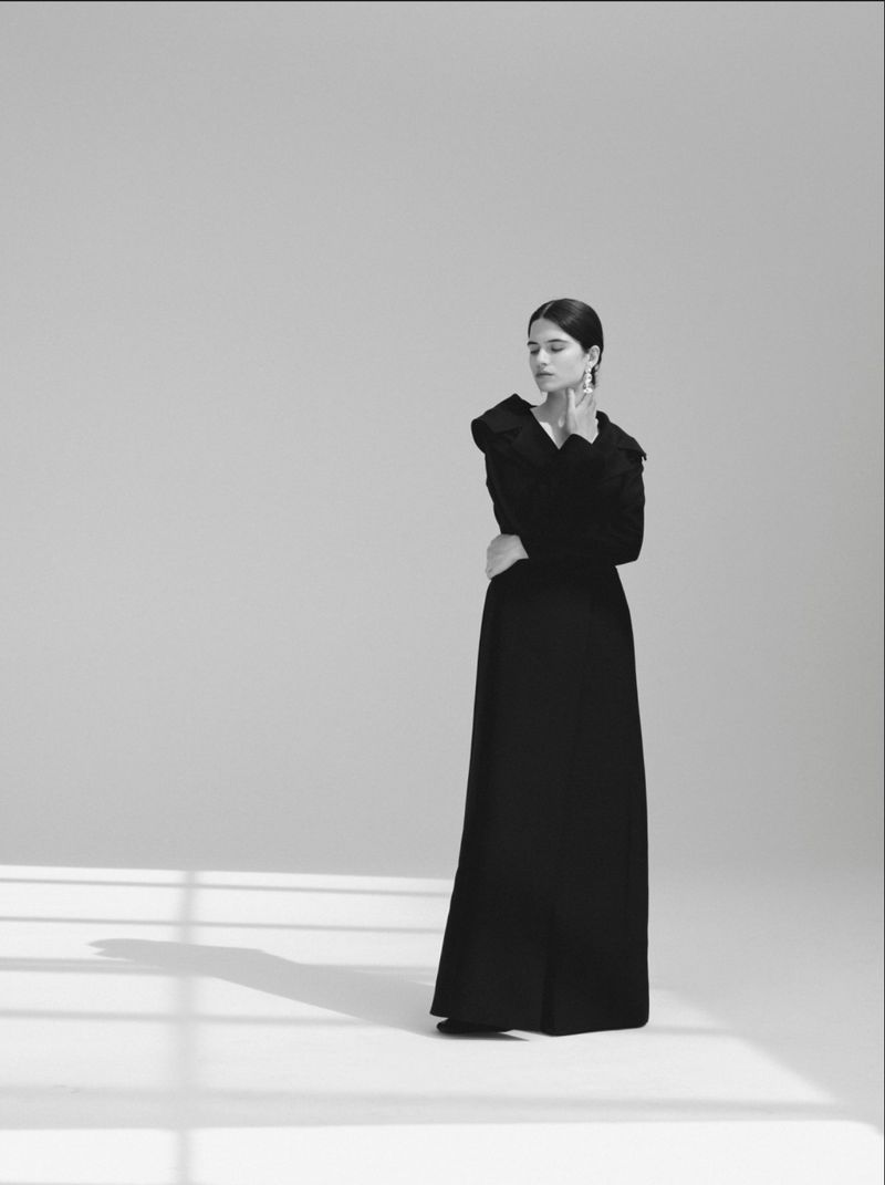 Model wearing a floor-length black dress standing in a studio with geometric window light cast across the floor.