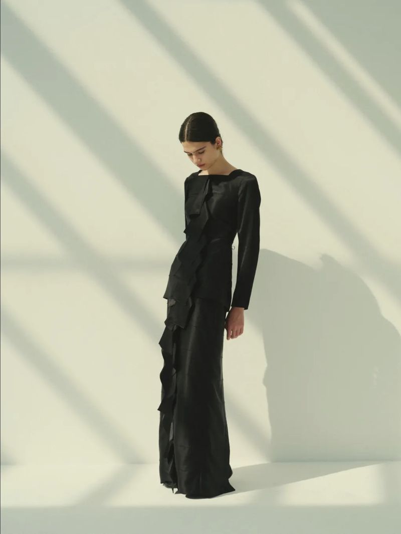 Model Peyton Cole posing in a full-length black long-sleeved dress with ruffle detailing against a pale backdrop with diagonal shadows.