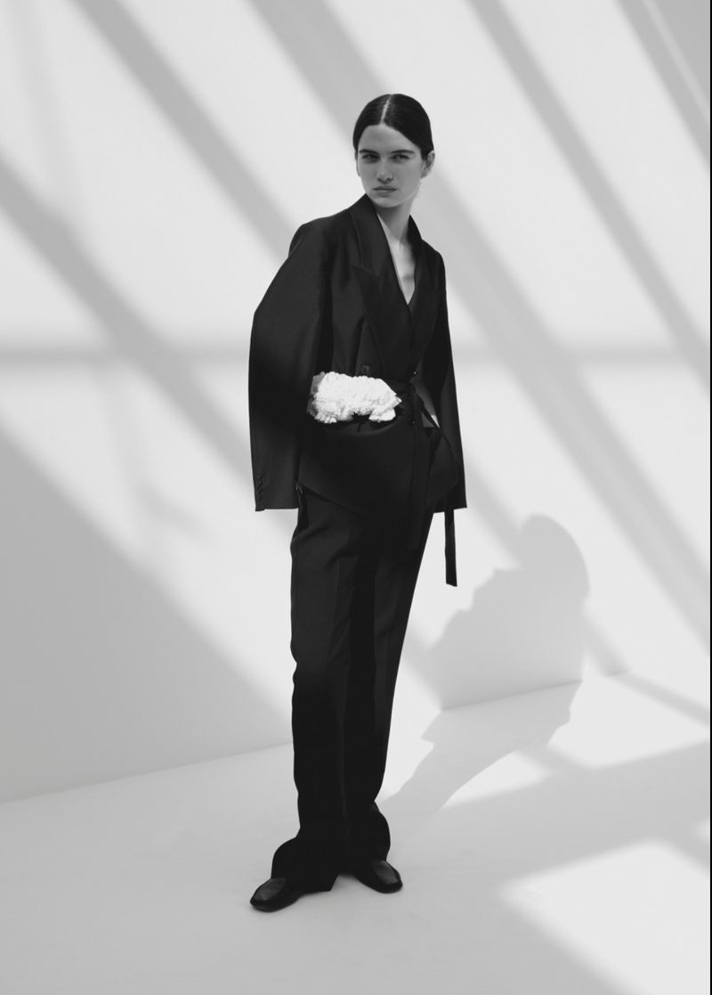 Model Peyton Cole standing in a black suit with a white ruffled waist detail against a white studio wall with diagonal shadows.