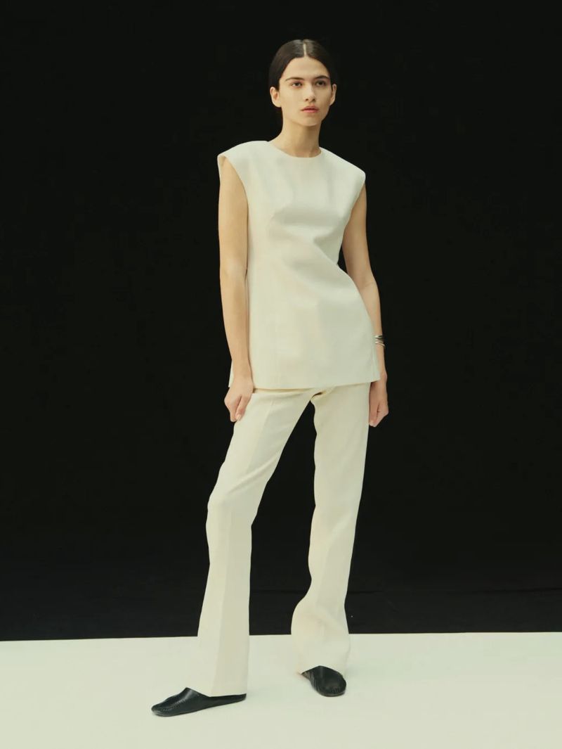 Model Peyton Cole wearing an off-white sleeveless structured tunic and matching tailored trousers against a black studio backdrop.