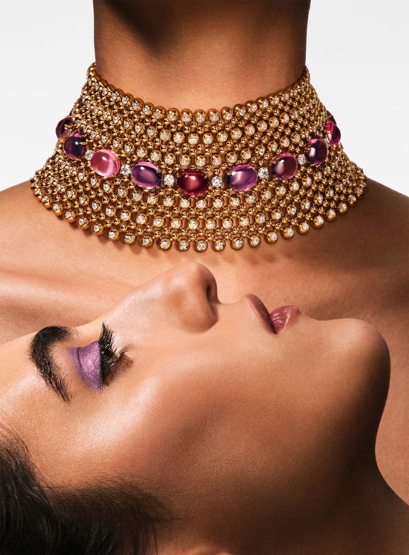 Close-up of a model wearing a wide gold choker necklace adorned with diamonds and pink cabochon gems beside another model in profile.