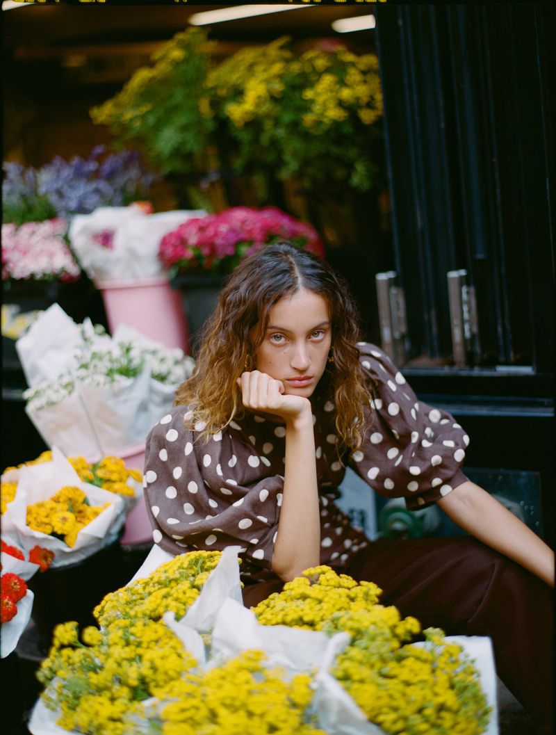 Model Sam Nixon seated in a brown polka dot puff-sleeve top surrounded by wrapped yellow flower bouquets at a market.