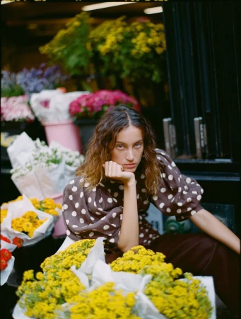 Model Sam Nixon seated in a brown polka dot puff-sleeve top surrounded by wrapped yellow flower bouquets at a market.