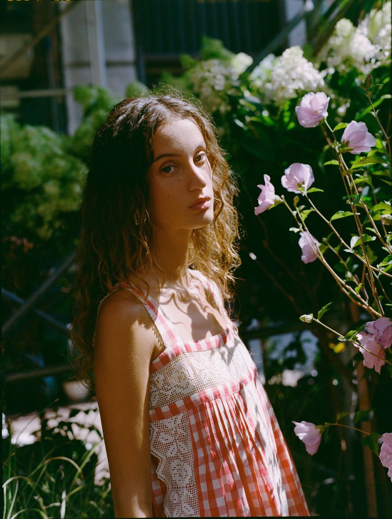 Model Sam Nixon wearing a pink and white gingham check dress with lace trim in a sunlit garden beside flowering pink blossoms.