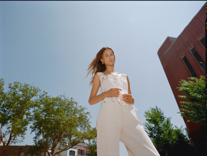 Low angle portrait of a woman in a white lace sleeveless top and cream trousers against trees, a brick building, and a blue sky.