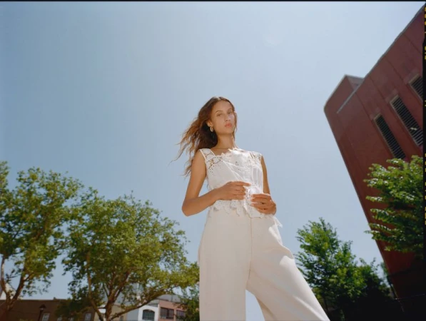 Low angle portrait of a woman in a white lace sleeveless top and cream trousers against trees, a brick building, and a blue sky.