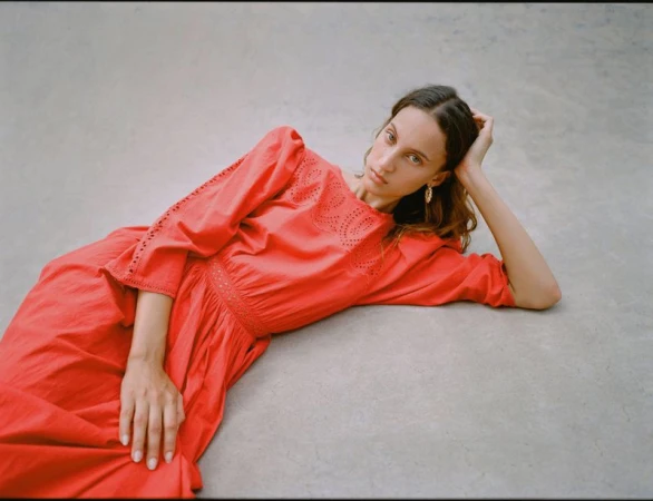 Model Sam Nixon reclining on a grey concrete surface wearing a bright coral red eyelet-detailed dress for Loeffler Randall.
