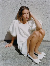 A woman crouching against a textured stucco wall wearing a white puff-sleeve mini dress and pointed white flats with large bows.