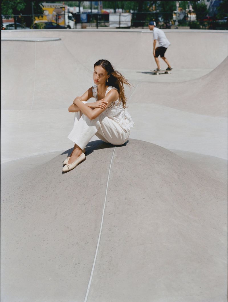 Model seated on a concrete skatepark mound wearing a white lace top, wide-leg trousers, and ballet flats with a skateboarder in the background.