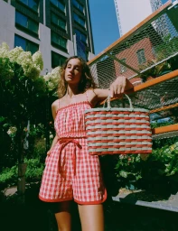 Model Sam Nixon wearing a red and white gingham two-piece set and holding a woven basket tote outdoors in direct sunlight.