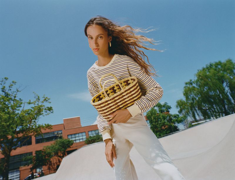 Model holding a woven basket tote bag wearing a striped long-sleeve top and cream trousers outdoors under a blue sky.