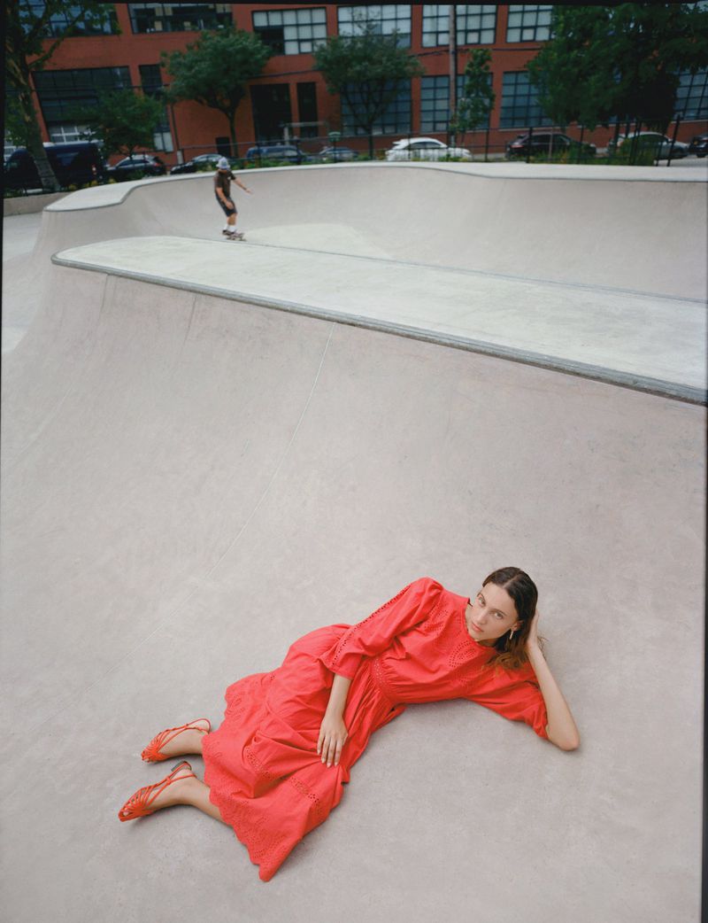 Model Sam Nixon in a vibrant coral-red dress and matching woven sandals reclining on a concrete skatepark bowl.