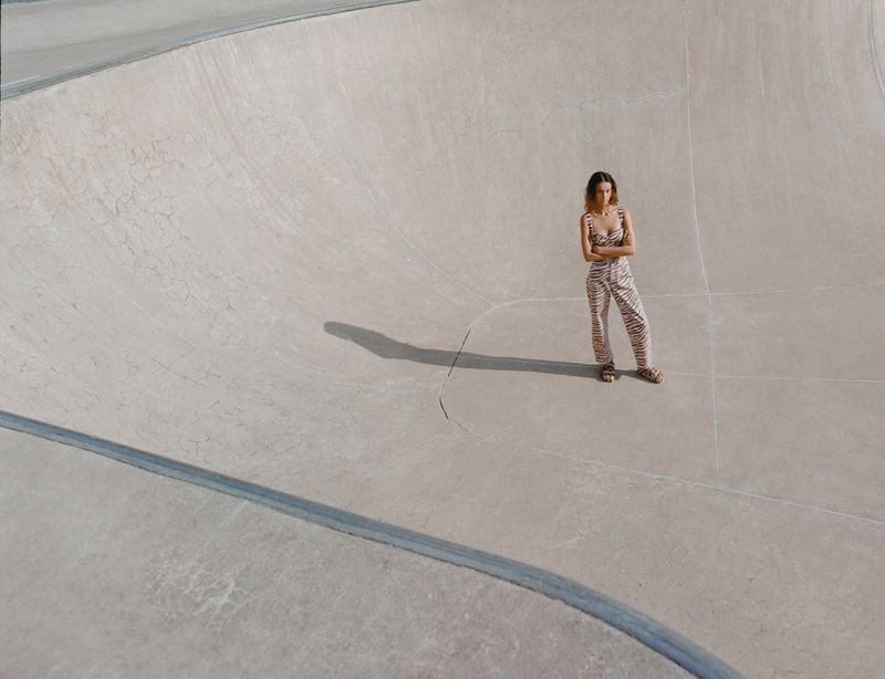 Model Sam Nixon stands with arms crossed wearing a patterned jumpsuit in an empty concrete skatepark bowl under direct sunlight.