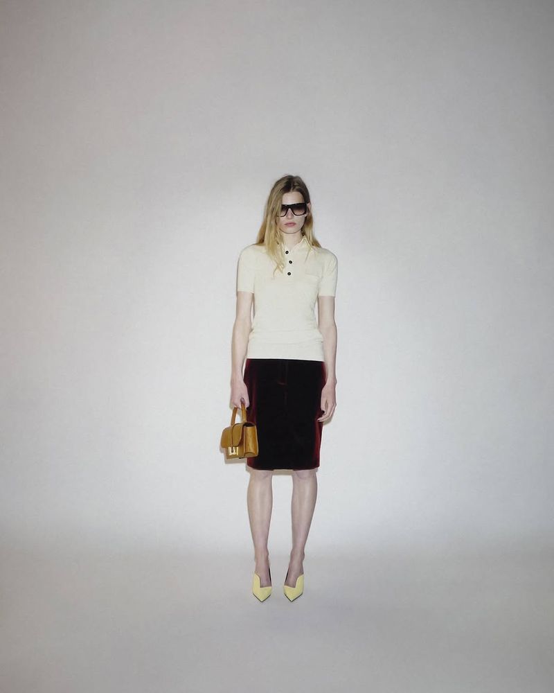 Model wearing a cream short-sleeve polo shirt, dark burgundy knee-length skirt, and yellow pumps against a studio background.