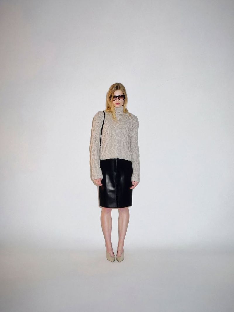 Model wearing a beige cable-knit turtleneck sweater, black leather pencil skirt, and dark sunglasses against a plain white backdrop.