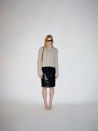 Model wearing a beige cable-knit turtleneck sweater, black leather pencil skirt, and dark sunglasses against a plain white backdrop.